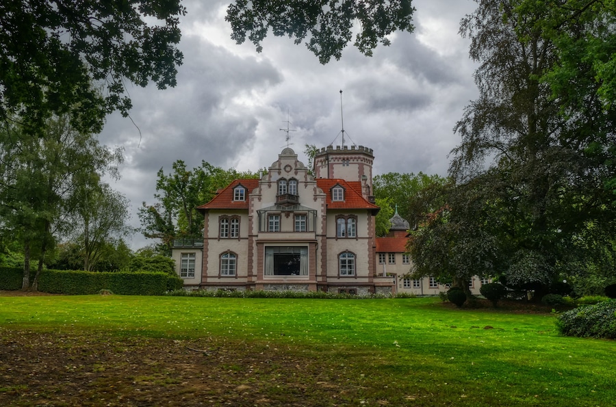 Un edificio grande e histórico con tejado de tejas rojas y una torre, rodeado de vegetación exuberante y árboles.