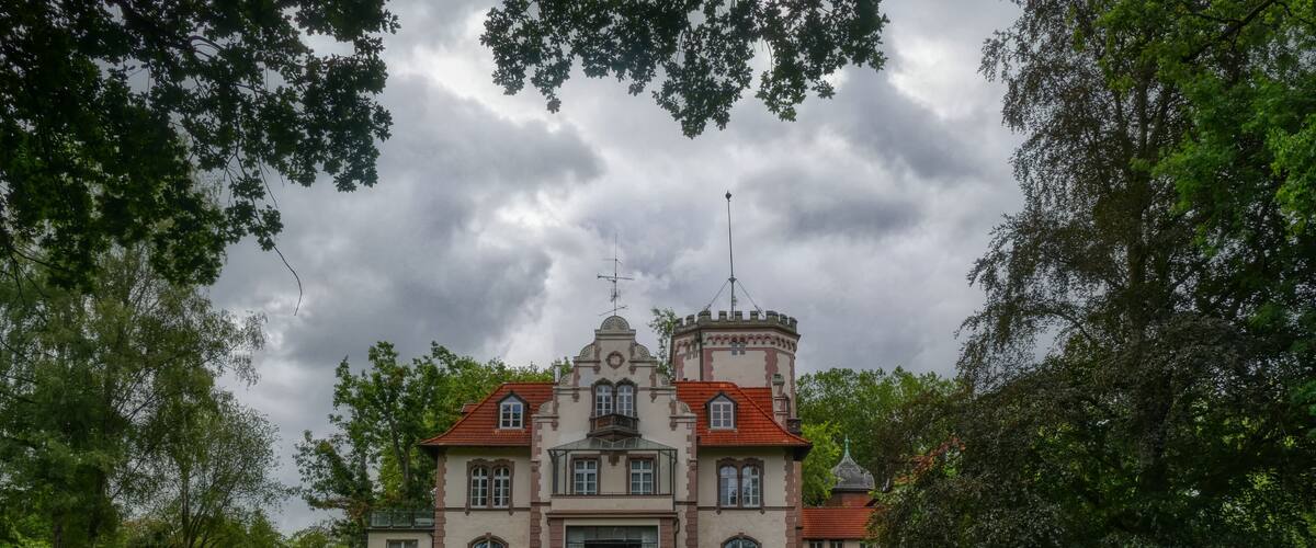 A large, historic building with a red-tiled roof and a tower, surrounded by lush greenery and trees.