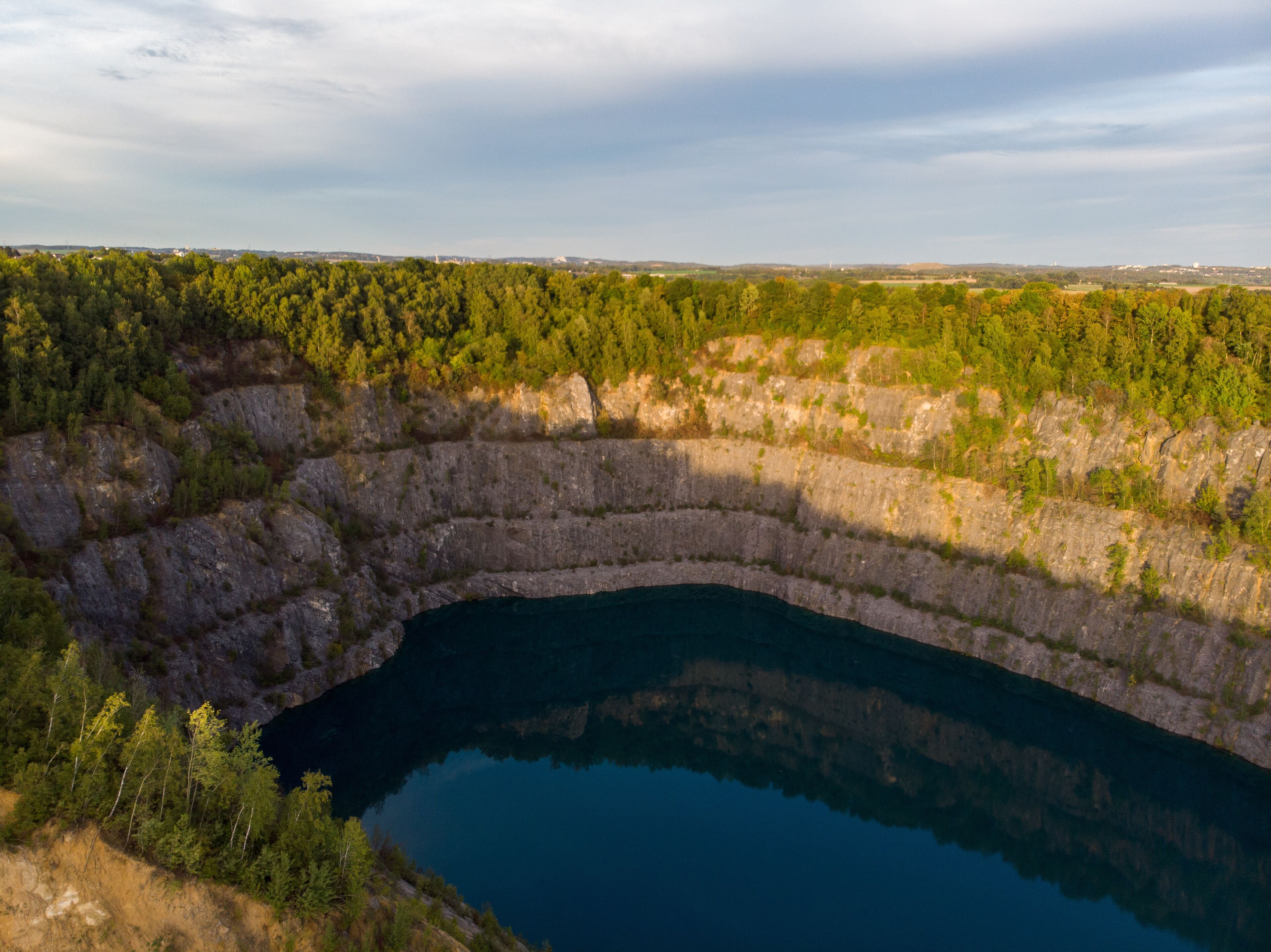 (Laufaufnahme, Drohne) Felswand eines Steinbruchs mit blauem See im Vordergrund