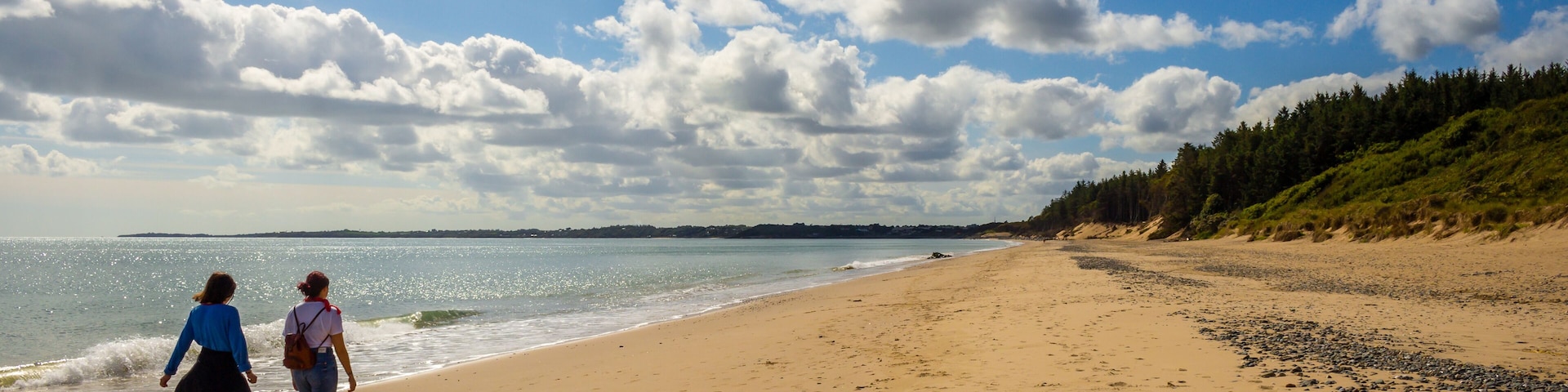 A sandy beach with footprints, a clear sky, and a forested hillside.