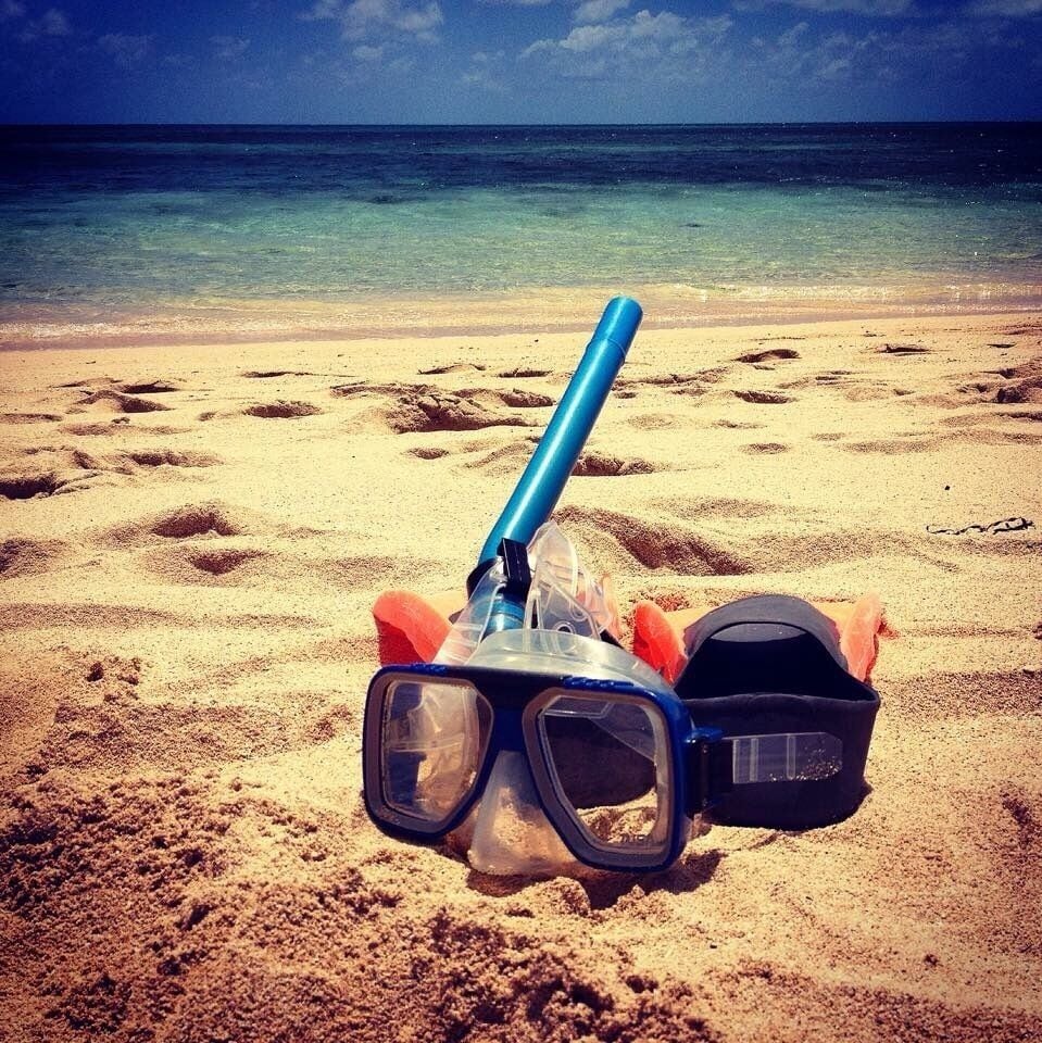 Una persona recostada en una playa arenosa, con un snorkel y antifaz azules, y una bolsa negra cerca.