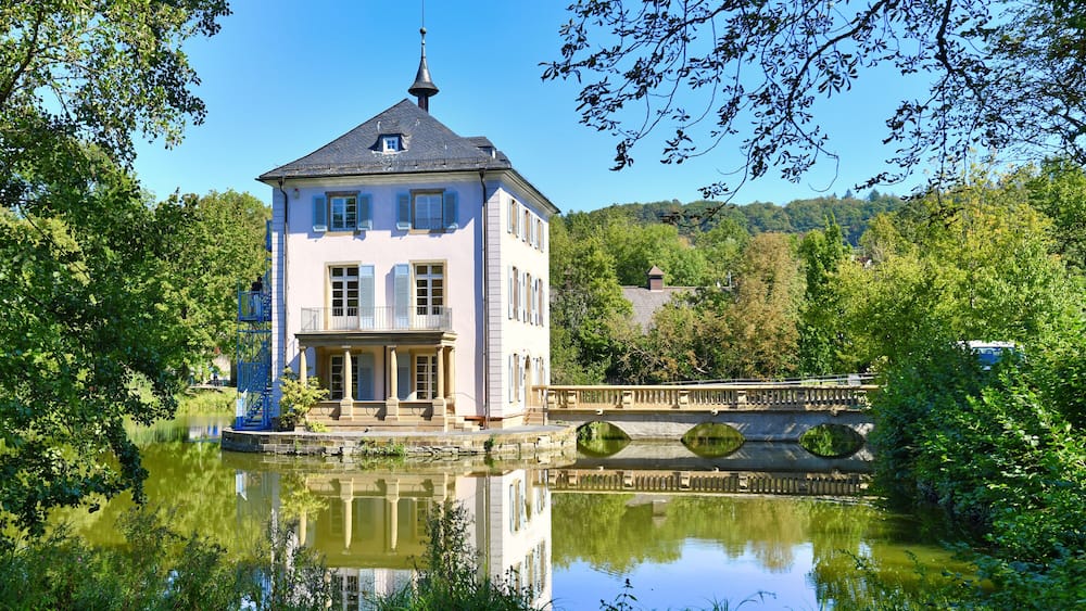 A stately building with a tower, a bridge over a calm river, and lush greenery surrounding the area.