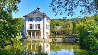 A stately building with a tower, a bridge over a calm river, and lush greenery surrounding the area.
