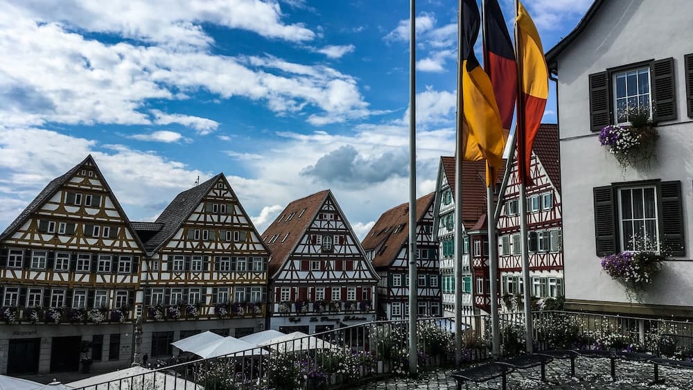 A cobblestone square with traditional half-timbered houses, a flagpole with multiple flags, and a building with flower boxes.