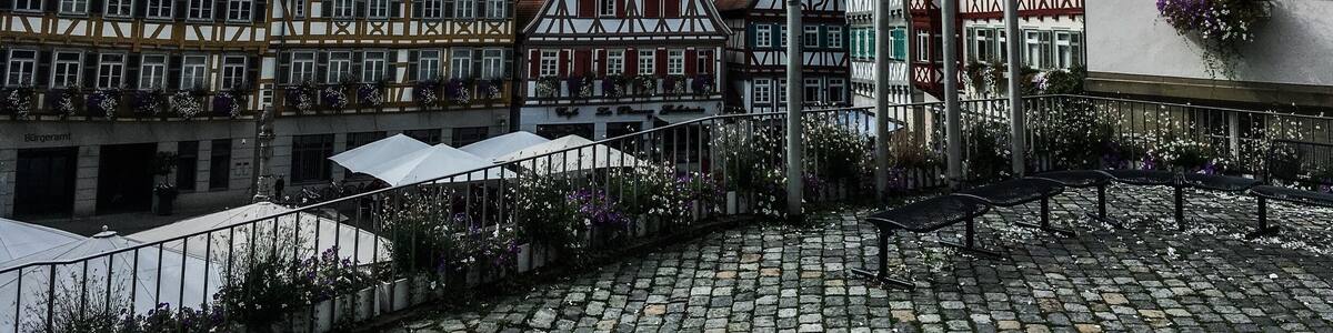 A cobblestone square with traditional half-timbered houses, a flagpole with multiple flags, and a building with flower boxes.