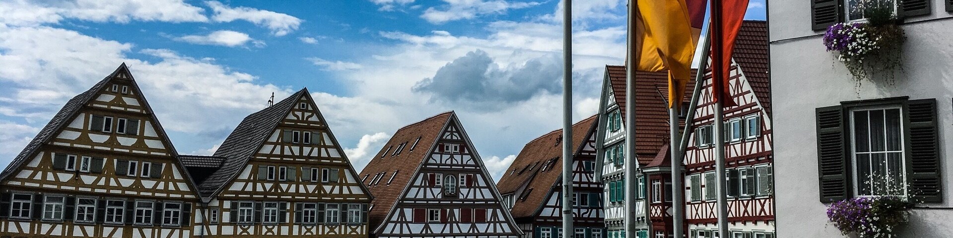 Uma praça de paralelepípedos com casas tradicionais em estilo enxaimel, um mastro com várias bandeiras e um edifício com caixas de flores.