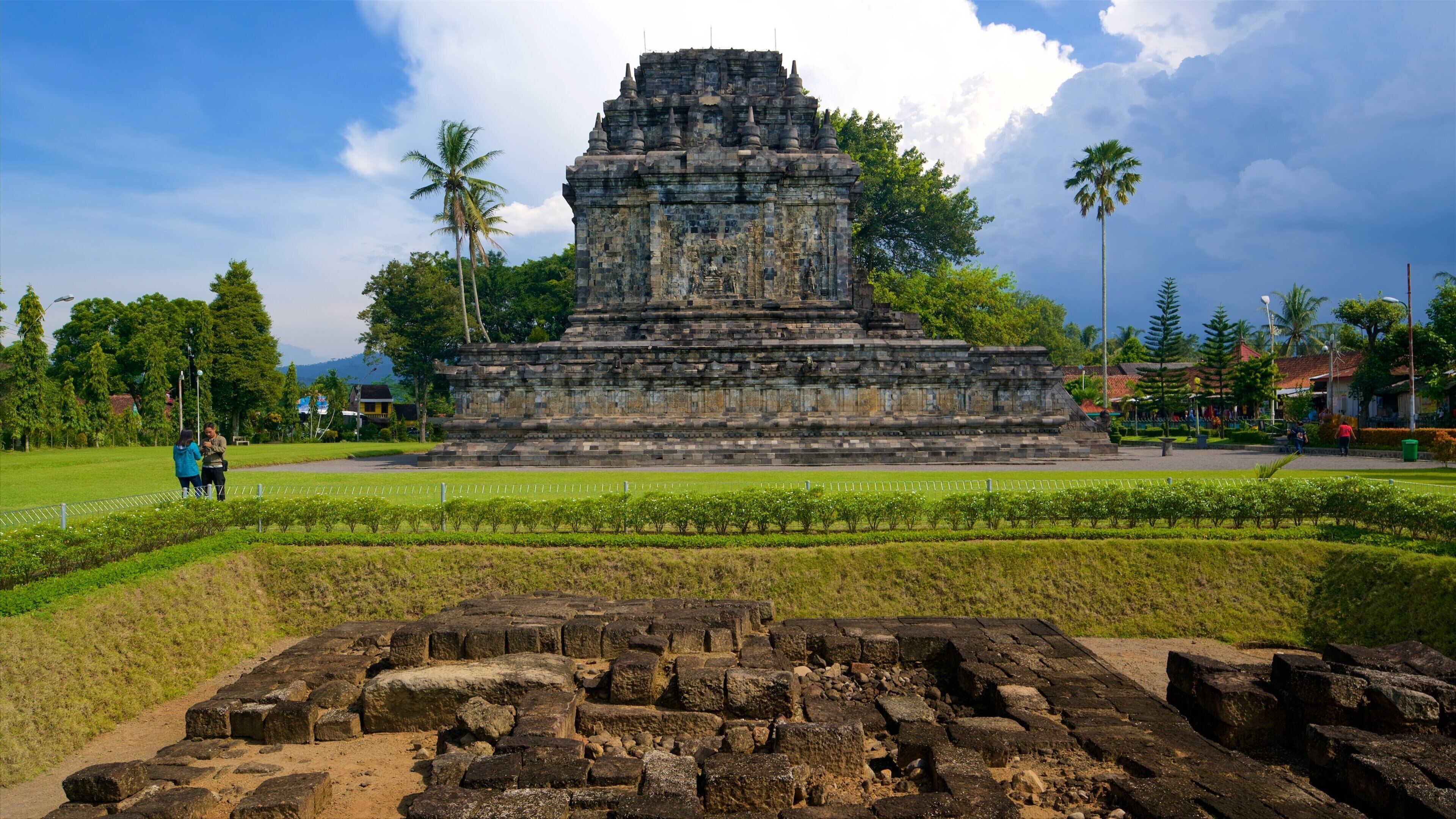 An ancient stone temple with a tiered structure, surrounded by lush greenery and palm trees.