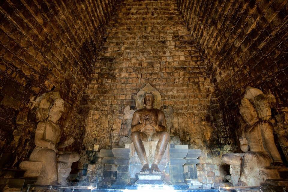 Eine große, sitzende Buddha-Statue in einem Tempel mit Ziegeldecke und Steinmetzarbeiten.