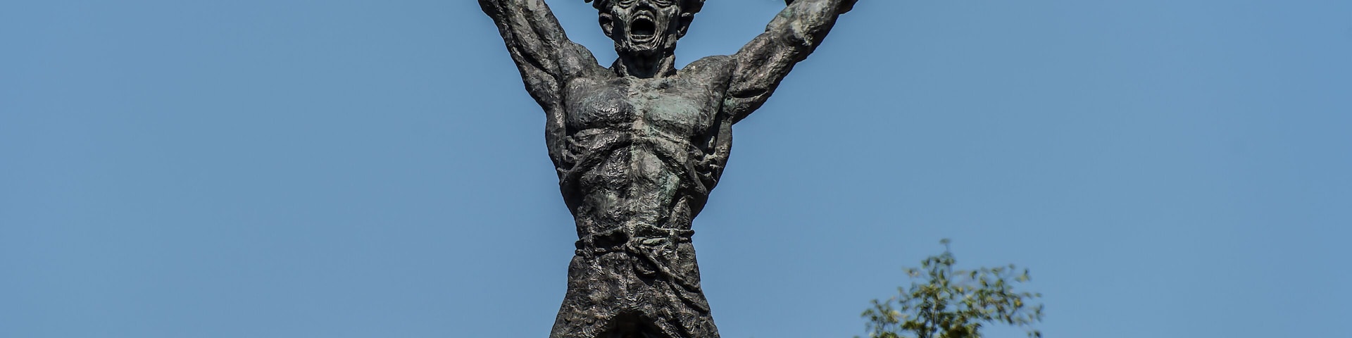 Eine bronzene Statue einer muskulösen Figur mit erhobenen Armen vor blauem Himmel, umgeben von Bäumen.