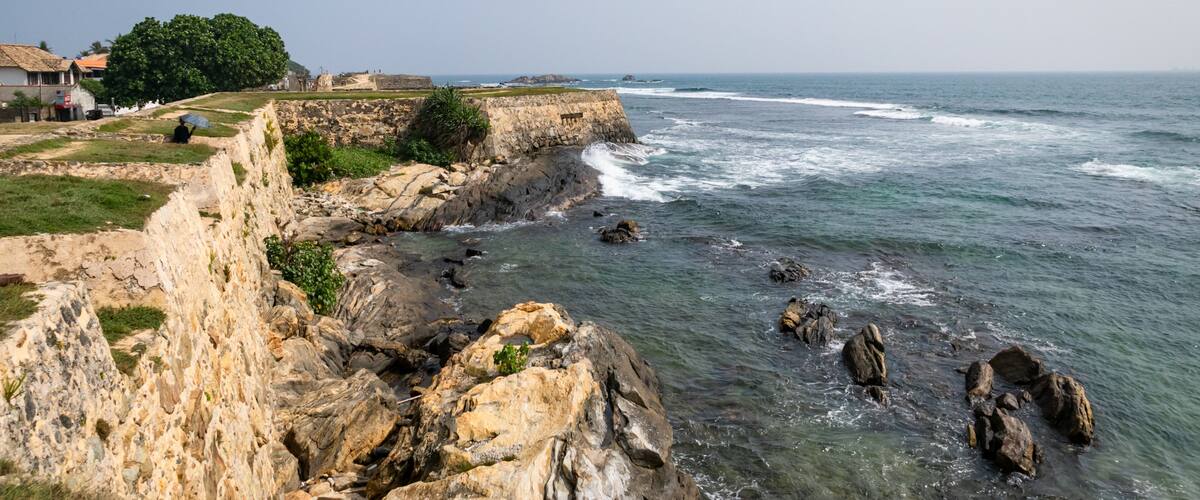 A coastal landscape with rocky cliffs, clear blue sea, and a few buildings in the background.