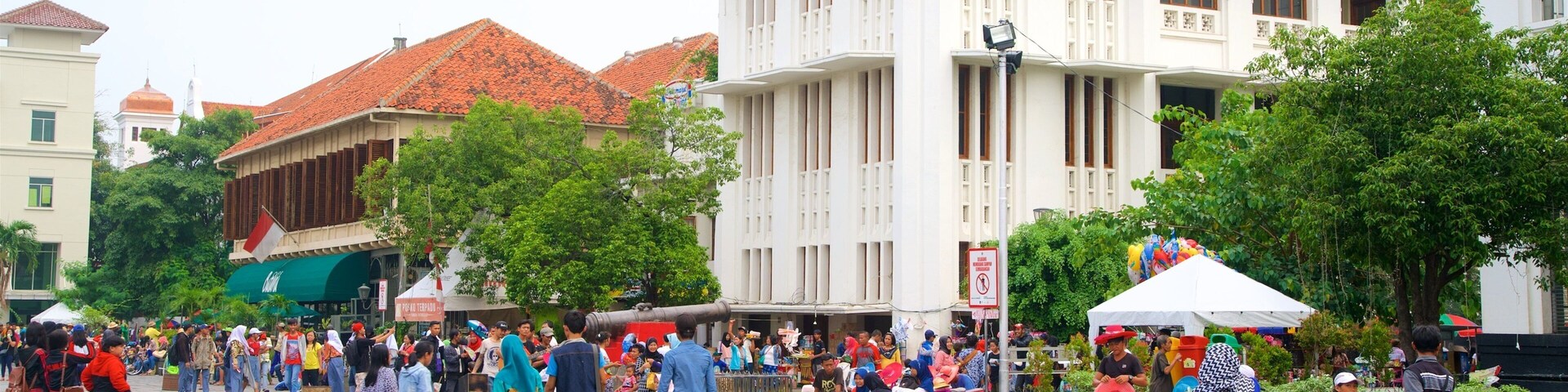 Una plaza pública concurrida, con gente caminando y sentada, rodeada de edificios con techos de tejas rojas y fachadas blancas.