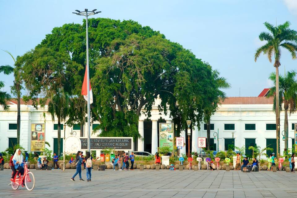 Una plaza pública con un árbol grande, un asta de bandera y un edificio con un cartel que dice "Museum Seni Rupa dan Keramik".