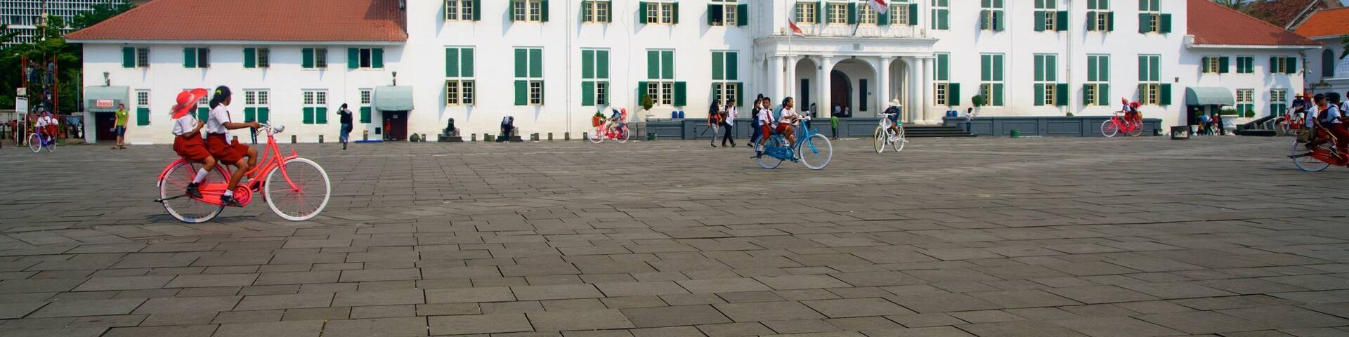 A large white building with green shutters and a red roof, surrounded by a paved square with people and bicycles.