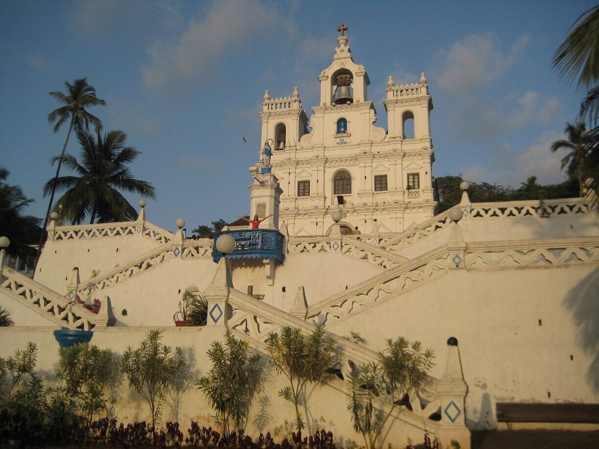 A white colonial-style church with a bell tower, surrounded by palm trees and a crowd of people.