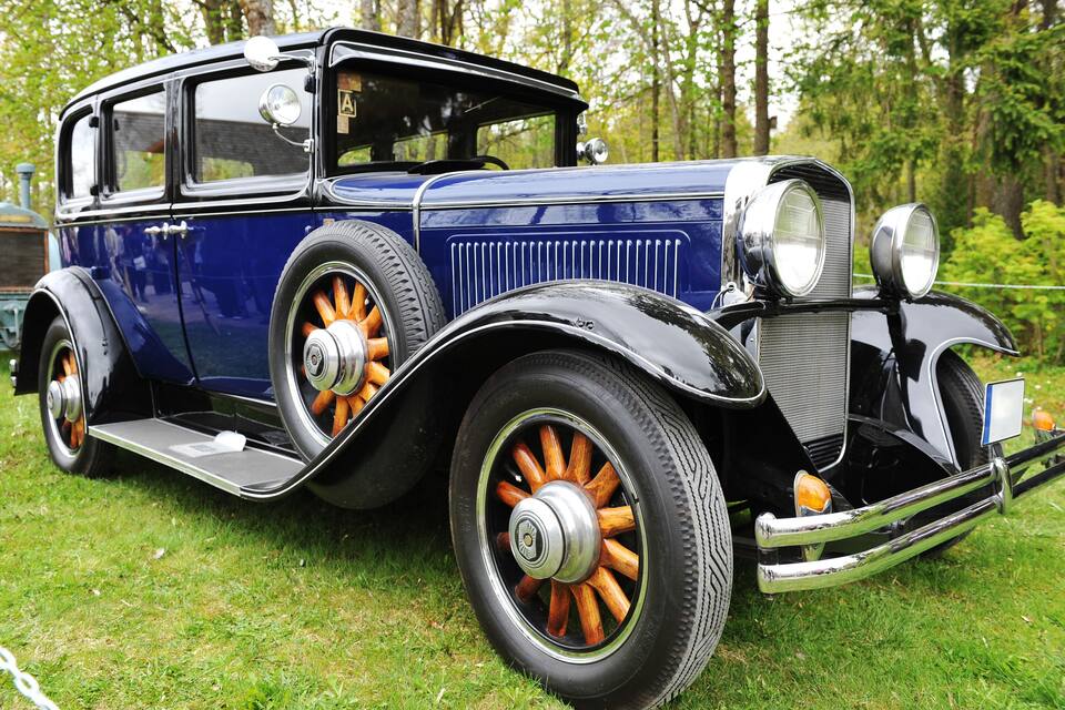 Une voiture bleue ancienne, avec des roues à rayons en bois et des détails chromés, est garée sur l’herbe.