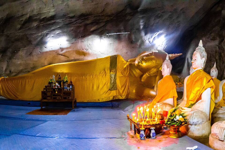 En hule med en liggende Buddha-statue, munke i gule klæder og et lille alter med stearinlys og blomster.