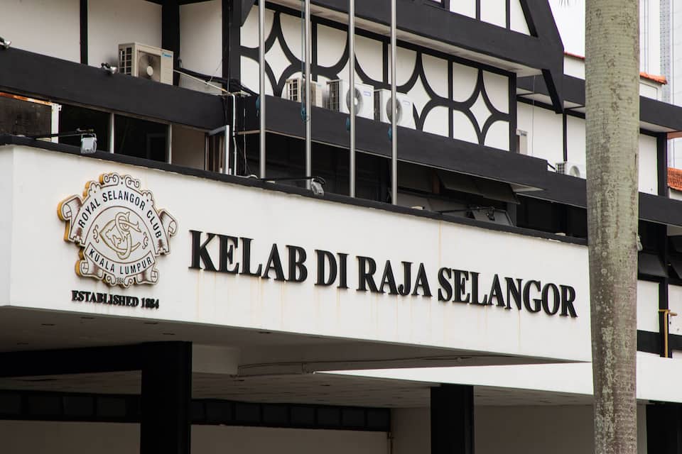 Rakennuksessa on kyltti, jossa lukee ”KELAB DI RAJA SELANGOR”, ja siinä on myös vaakuna, jossa on teksti ”ROYAL SELANGOR CLUB KUALA LUMPUR, PERUSTETTU 1884”.