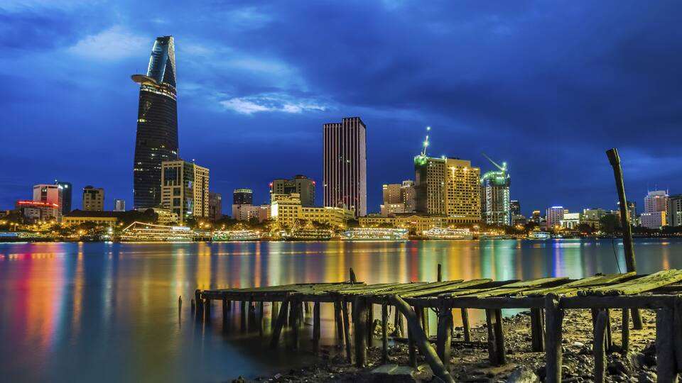 A nighttime city skyline with a prominent skyscraper reflecting on calm waters, a dock, and various illuminated buildings.