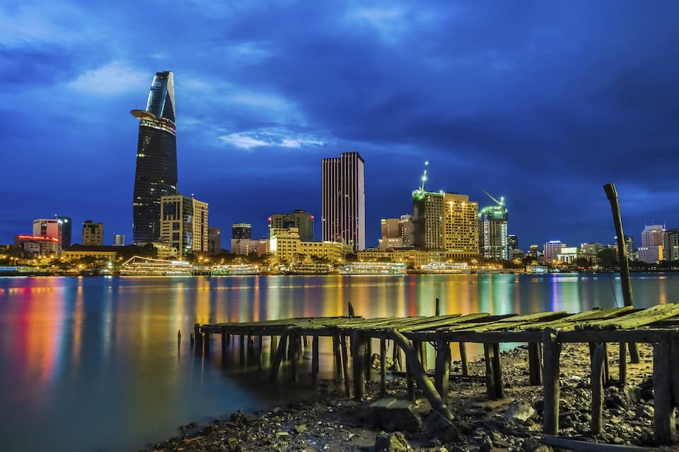 Un horizonte urbano nocturno con un rascacielos destacado reflejado en aguas tranquilas, un muelle y varios edificios iluminados.