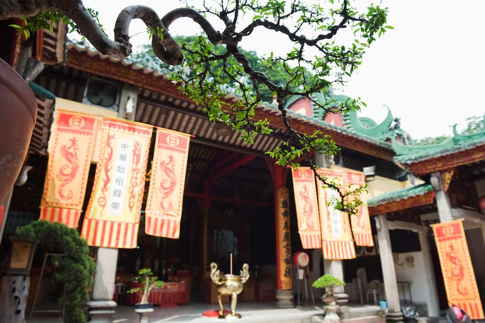 Ein traditioneller chinesischer Tempel mit aufwendiger Dekoration, darunter rote Banner mit chinesischen Schriftzeichen und ein goldener Weihrauchbrenner im Vordergrund.
