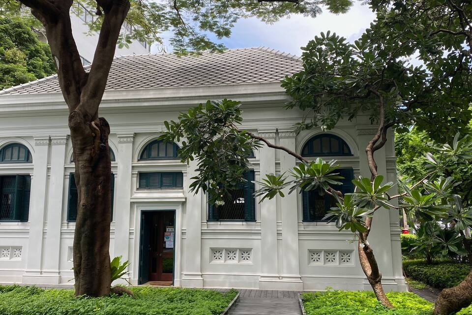 A white colonial-style building with arched windows and a tiled roof, surrounded by lush greenery.