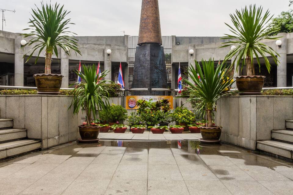 A plaza with a tall, pointed monument, surrounded by potted plants and flags.