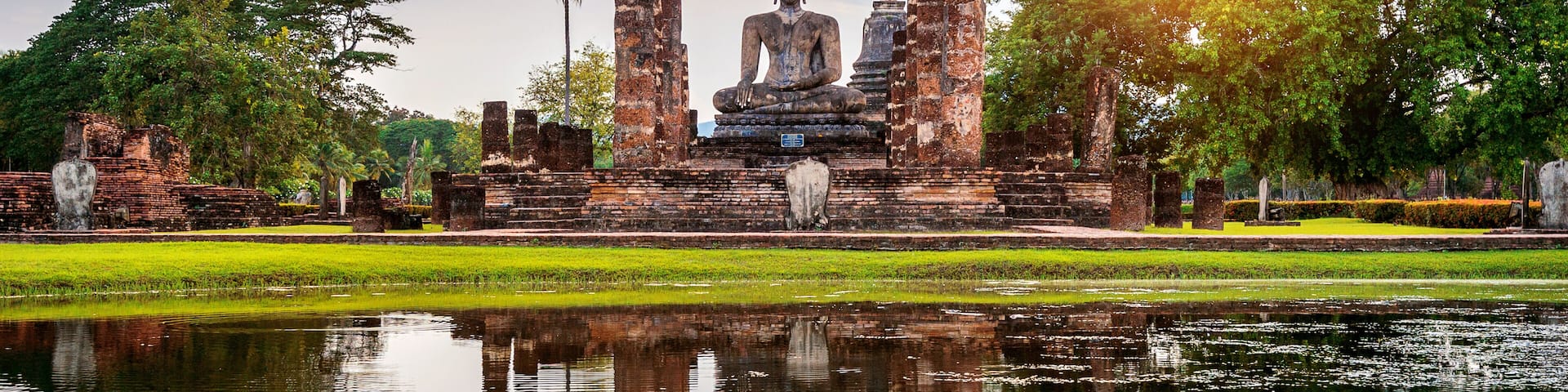 Una gran estatua de Buda rodeada de ruinas de ladrillo antiguo y un estanque tranquilo que refleja la estatua.