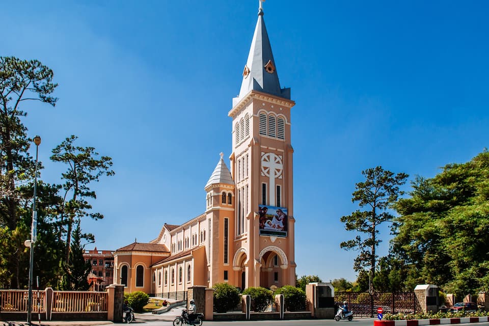 A church with a tall tower and a large cross, surrounded by trees and a clear blue sky.