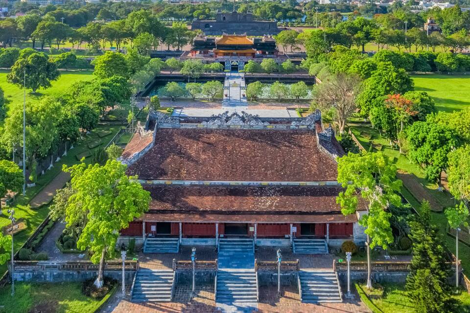 Um edifício tradicional com telhado em telhas, rodeado por árvores verdes e um céu azul límpido.