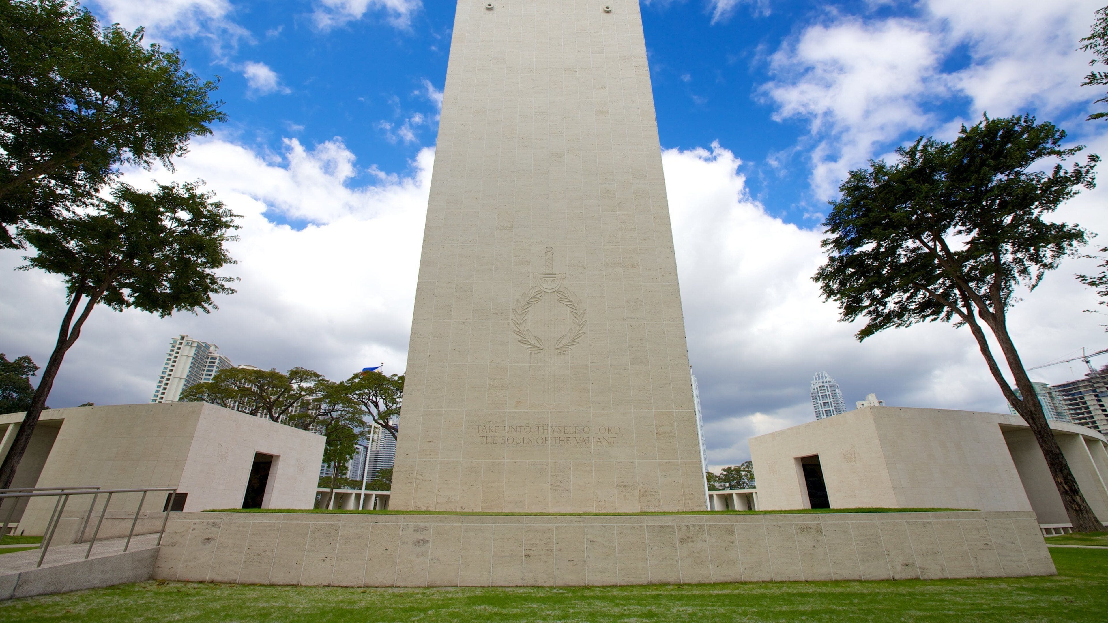A tall monument with an inscription, surrounded by grass and trees, with buildings in the background.