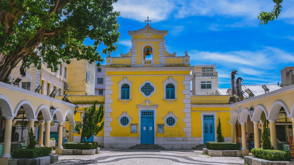 A yellow church with a blue door and arches, surrounded by cobblestone pavement and greenery.