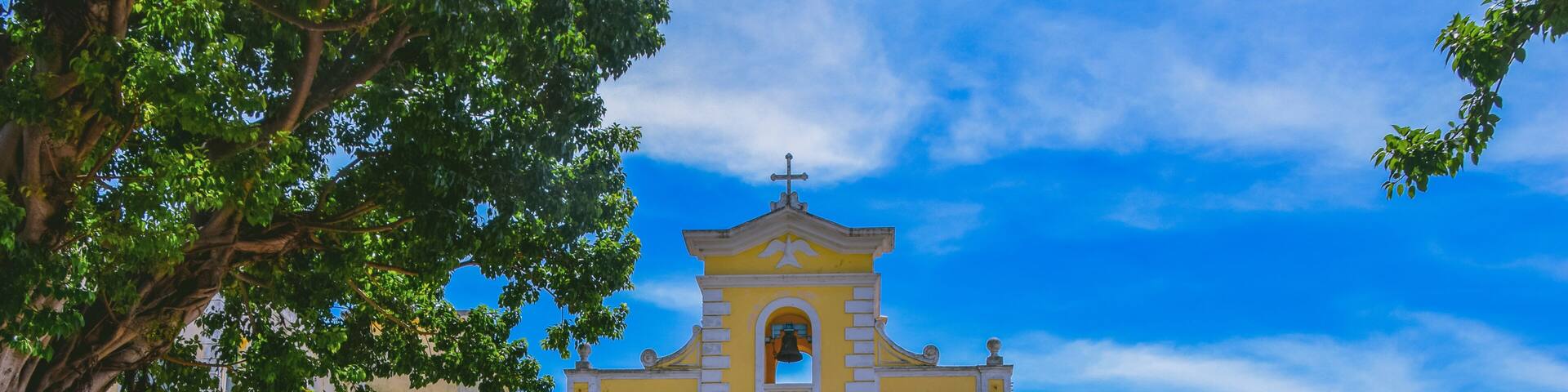A yellow church with a blue door and arches, surrounded by cobblestone pavement and greenery.