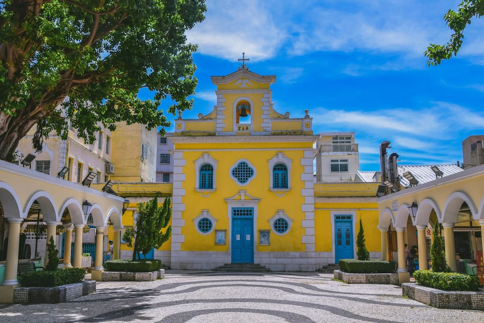 Una iglesia amarilla con puerta azul y arcos, rodeada de adoquines y vegetación.