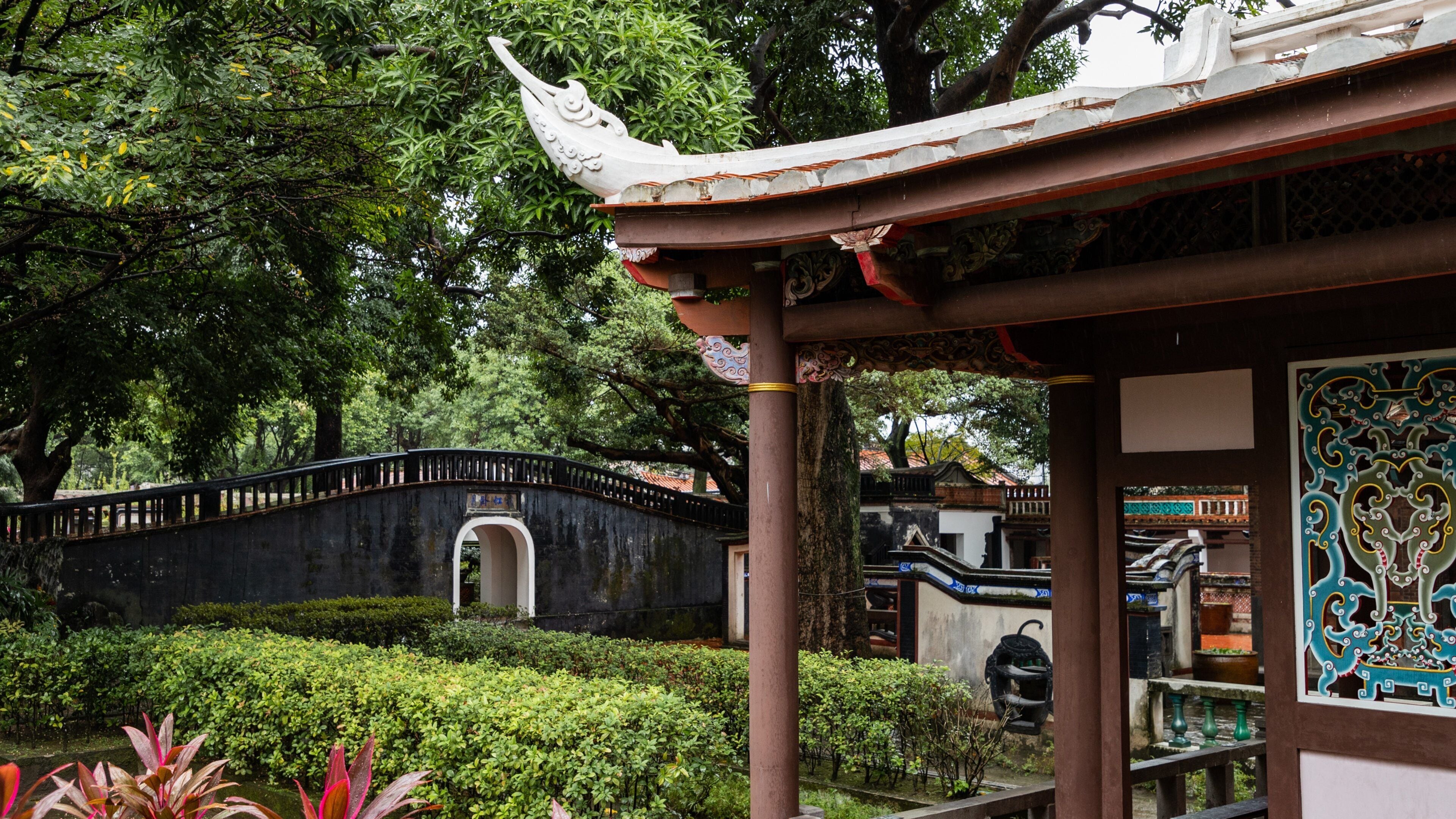 A traditional Chinese pavilion with ornate roof tiles, surrounded by lush greenery and a stone bridge.