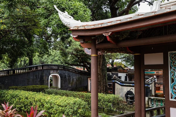 Un pavillon chinois traditionnel, orné de tuiles décoratives sur le toit, entouré d’une végétation luxuriante et surplombant un pont de pierre.
