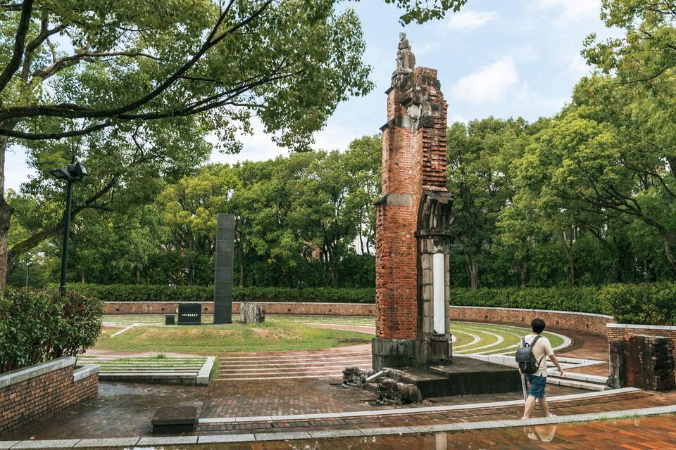 Un monumento histórico con una torre de ladrillo y una estatua, rodeado de exuberante vegetación y un sendero pavimentado.