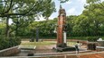 A historical monument with a brick tower and a statue, surrounded by lush greenery and a paved walkway.