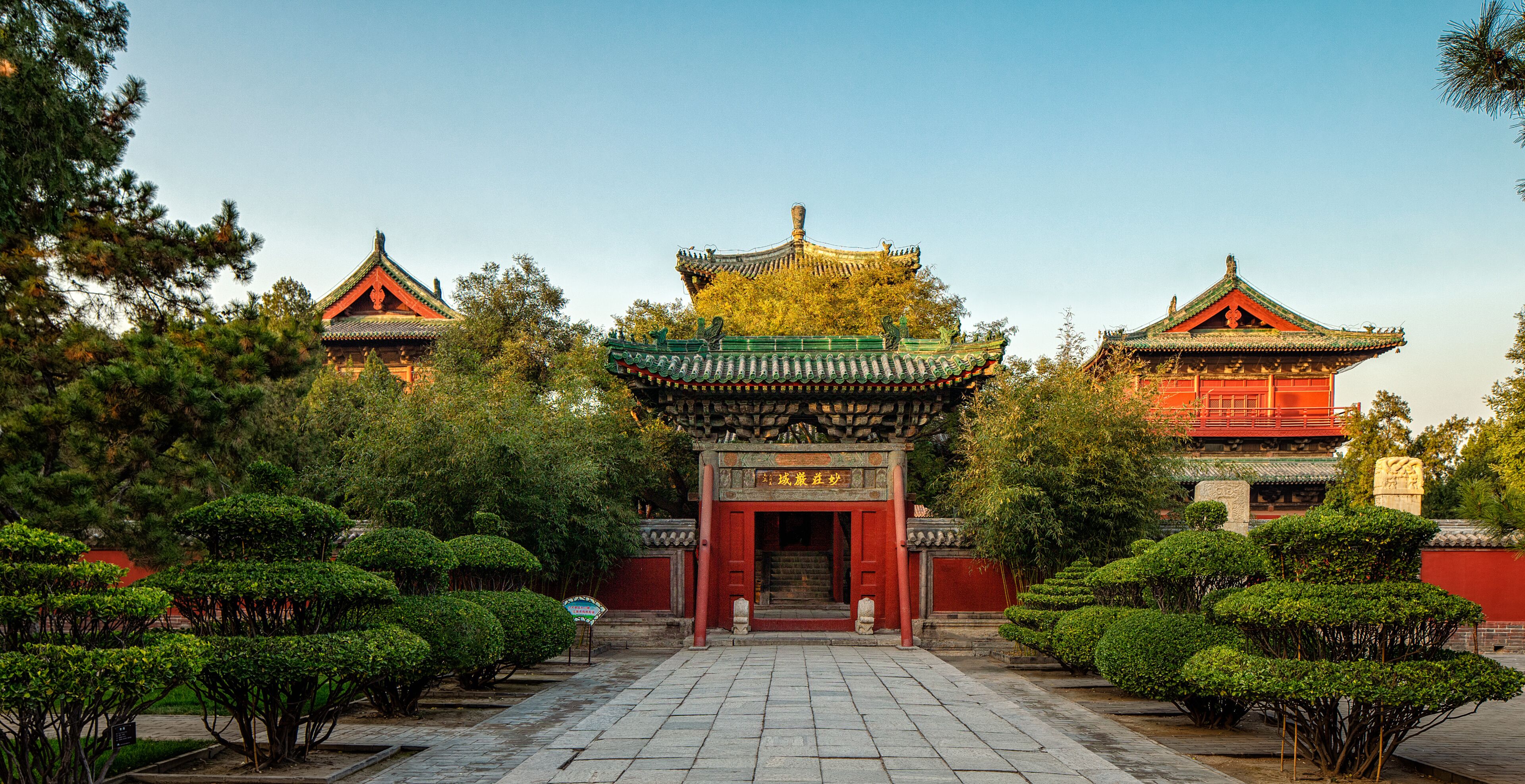 A traditional Chinese gate with ornate architecture, surrounded by well-maintained trees and a clear sky.