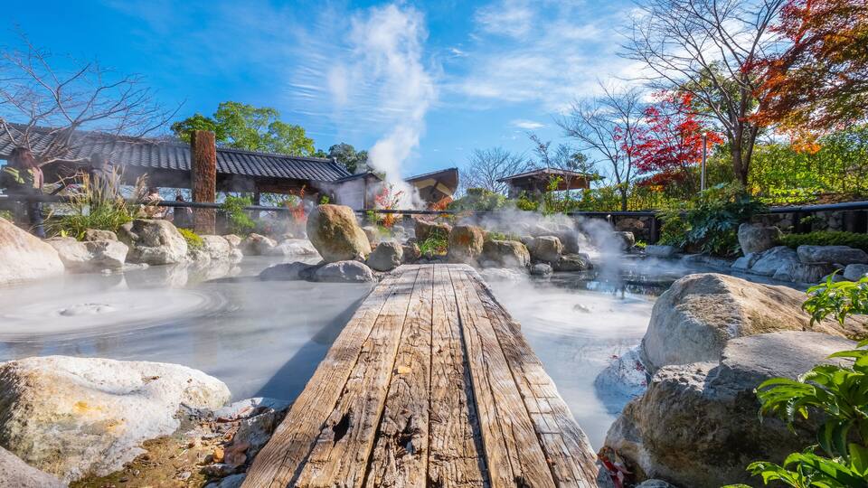 A wooden boardwalk leading to a hot spring area with steam rising from the water, surrounded by rocks and trees with autumn foliage.