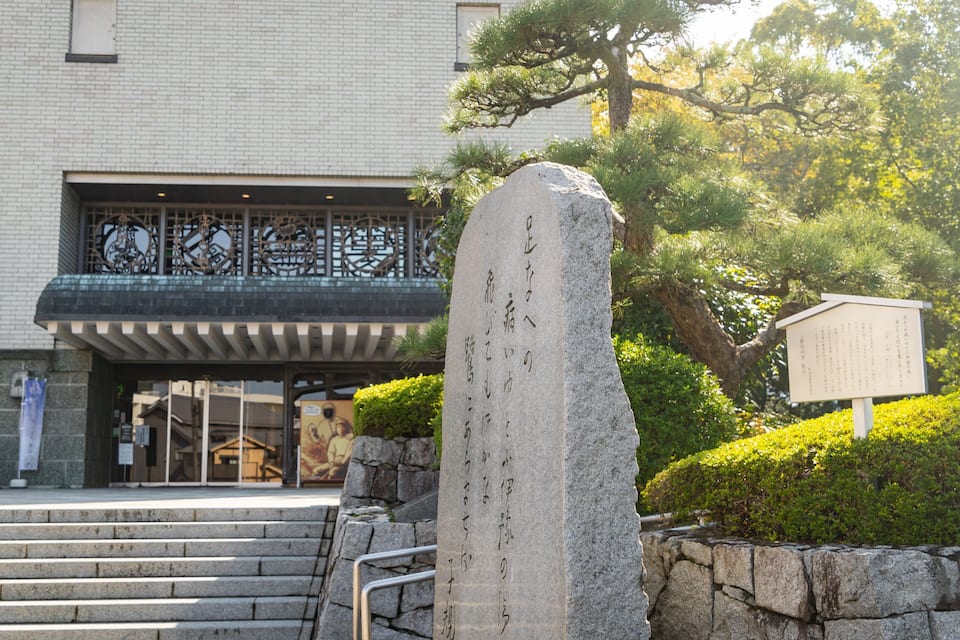 Un monument de pierre orné d’inscriptions en japonais, une entrée de bâtiment surmontée d’une porte traditionnelle, et des marches menant à l’entrée.