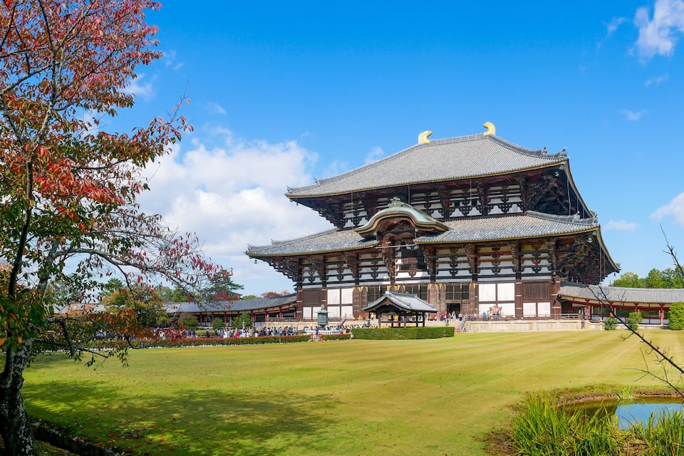 A traditional Japanese building with a large, multi-tiered roof and wooden architecture, surrounded by a well-maintained lawn and a small pond.