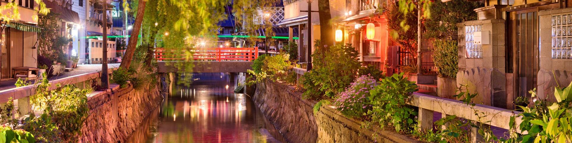 A serene riverside scene with traditional architecture, a wooden bridge, and lush greenery.