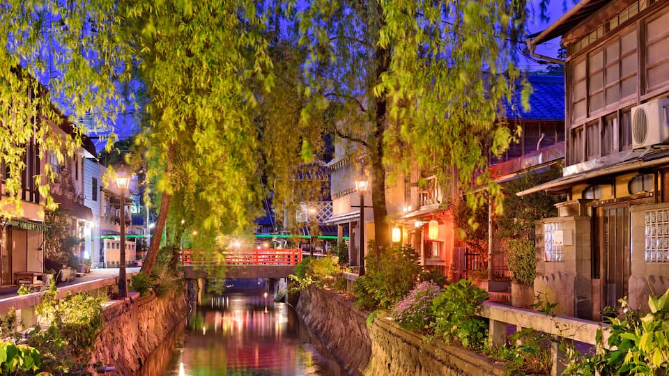 A serene riverside scene with traditional architecture, a wooden bridge, and lush greenery.