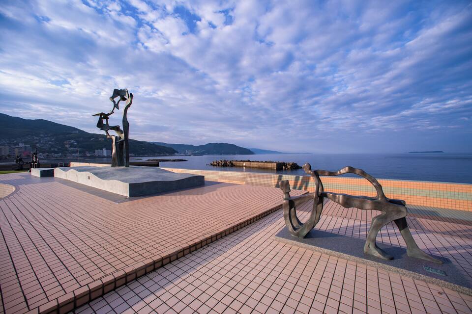 A waterfront park with a sculpture, a tiled walkway, and a distant view of a city and mountains.