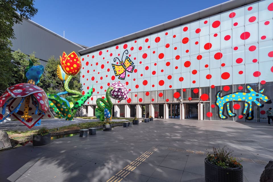 Um edifício com fachada em vermelho e branco com bolinhas, esculturas coloridas grandes e uma escultura de animal em azul e amarelo.