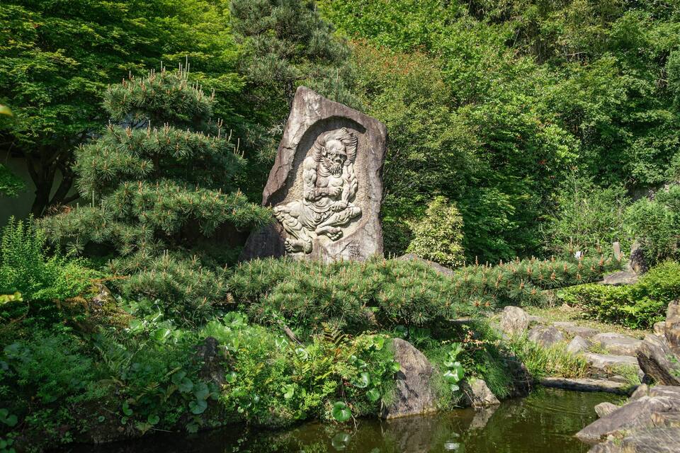 Scultura in pietra raffigurante una figura in un ambiente naturale, con vegetazione lussureggiante e uno stagno.