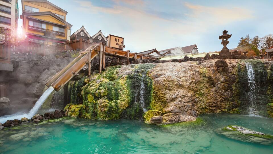 A traditional Japanese onsen with a wooden staircase leading to a hot spring, surrounded by buildings and a stone lantern.