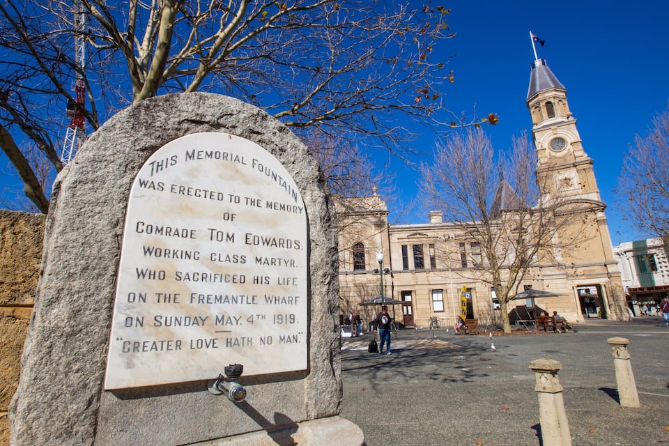 A memorial fountain with a plaque, a clock tower, and a building in the background.