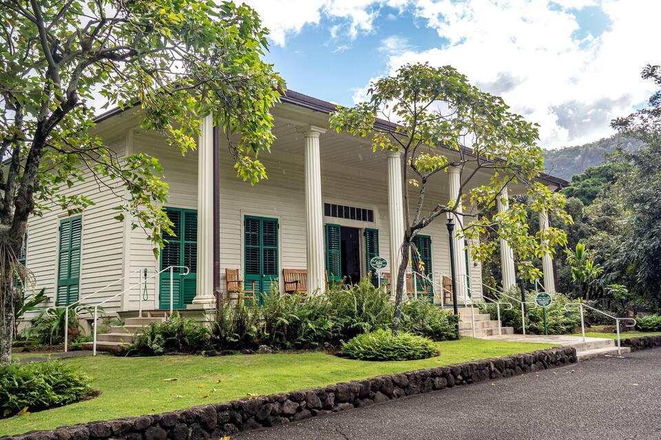 Une maison de style colonial, aux murs blancs et aux volets verts, entourée d’une végétation luxuriante et d’un mur de pierre.