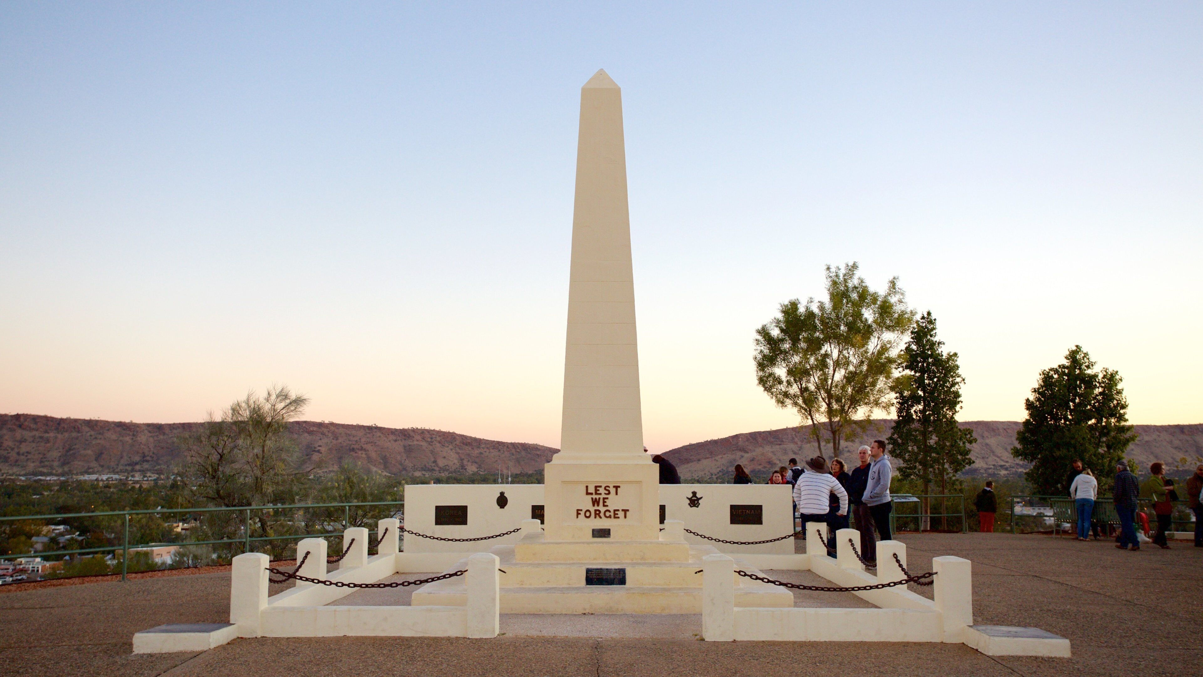 Et højt, hvidt obelisk med teksten 'Lest We Forget' indgraveret ved foden, omgivet af et pigtrådshegn, foran en bakke og træer.